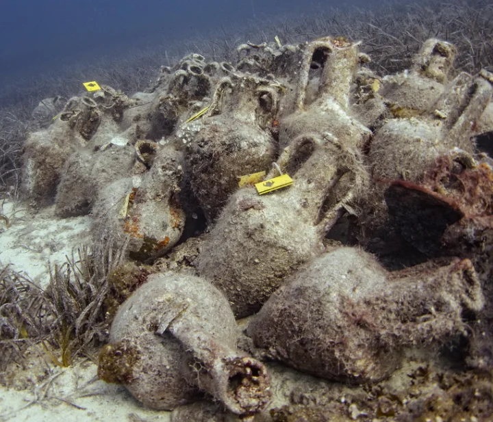 Stacked ancient amphorae from the Peristera shipwreck in Alonnisos, Greece, are partially buried on the seabed with marine growth and sediment.